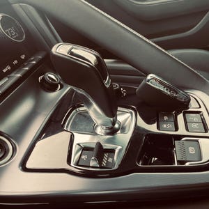 Puede incluir: Interior de coche con palanca de cambios, llave y panel de control. La consola es negra con detalles plateados, y una pantalla muestra una temperatura de 23 grados Celsius. Diseño elegante y moderno.