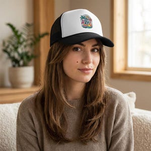 Puede incluir: Gorra de camionero en blanco y negro con un panel frontal blanco con un gráfico colorido de una escena tropical y el texto "Aloha Lil". La gorra tiene una visera curva y una parte trasera de malla.