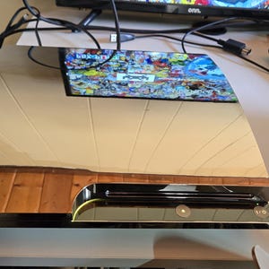 Puede incluir: Una consola de juegos plateada y negra con una superficie superior reflectante. La consola está sobre una superficie blanca con un borde de madera. Una pantalla se refleja en la superficie de la consola y un mando a distancia está cerca.