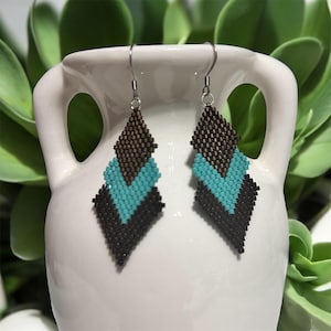 Può includere: Un paio di orecchini di perline dal design geometrico. Gli orecchini presentano una forma a diamante con una sezione superiore marrone, un motivo a V turchese e una sezione inferiore nera. Sono appesi a ganci color argento.
