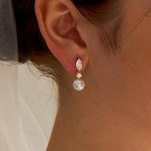 Könnte beinhalten: Elegante, goldfarbene Ohrhänger mit einem tropfenförmigen Edelstein und einer weißen Perle. Die Ohrringe haben ein zartes Design, ideal für Braut- oder formelle Anlässe.