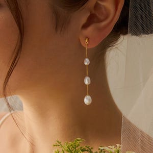 May include: Gold drop earrings with three white, teardrop-shaped pearls hanging from a fine chain. The earrings are displayed against a soft background, emphasising the jewellery's elegant design. The pearls have a lustrous sheen.