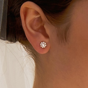 Könnte beinhalten: Ein goldfarbener Ohrstecker mit einer zentralen Perle, umgeben von kleinen, klaren Edelsteinen. Der Ohrring ist vor dem Hintergrund eines Ohrs und Haares platziert, was auf Schmuck und Accessoires hindeutet. Das Design ist blütenförmig.