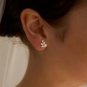 May include: A gold stud earring in a leaf design, set with clear, marquise-cut stones and a small, round pearl. The earring is displayed on an ear, highlighting its delicate features and elegant style.