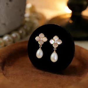 Peut inclure: Une paire de boucles d'oreilles dorées. Chaque boucle d'oreille présente un motif de fleur à quatre pétales avec un petit diamant au centre et une perle en forme de goutte. Les boucles d'oreilles sont présentées sur un support en velours noir.