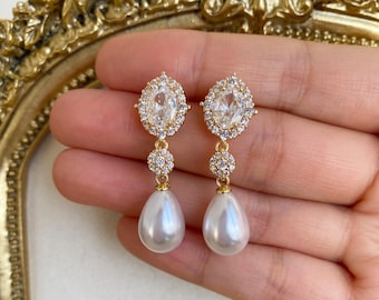 May include: A pair of gold-coloured drop earrings. Each earring has a large, oval clear stone surrounded by smaller clear stones. Below the oval stone is a cluster of smaller clear stones, and a pear-shaped white pearl hangs at the bottom.
