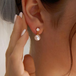 May include: A gold-colored earring with a small, clear gemstone at the top and a dangling white pearl. The earring is worn on the ear of a person. The person's skin is visible in the image.