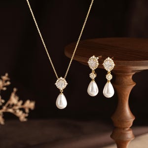 May include: A gold-toned jewellery set consisting of a necklace and a pair of drop earrings. Each piece features a central oval-shaped clear stone surrounded by small white crystals, with a smooth white teardrop-shaped pearl pendant hanging below.