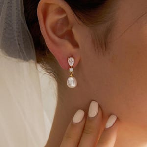Puede incluir: Elegantes pendientes dorados con una gota de perla blanca en forma de pera. Los pendientes presentan una piedra transparente en forma de pera en la parte superior y una pequeña piedra redonda en el centro. Se llevan en la oreja.