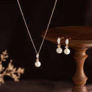 May include: A gold necklace and a pair of matching earrings, each featuring a white pearl suspended below a small round clear stone and a marquise-cut clear stone. The jewelry set is crafted with thin gold chains and gold metal settings.