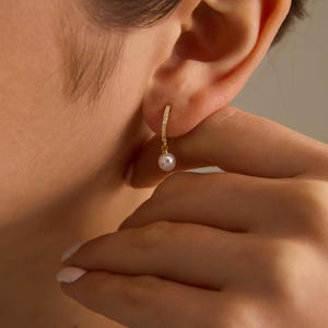 May include: A gold hoop earring with small, clear stones and a dangling white pearl. The earring is worn on an ear, with a hand gently touching the ear. The jewelry is simple and elegant.