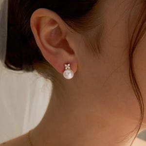 Può includere: Primo piano di un orecchino di perle con un design floreale color oro. L'orecchino presenta una grande perla bianca rotonda e un gruppo di piccole pietre scintillanti. L'orecchino è indossato su un orecchio.