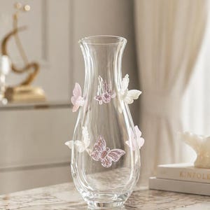 May include: A clear glass vase with a flared top and a rounded body. The vase is adorned with several decorative butterflies in shades of pink and white. The vase is sitting on a reflective surface.