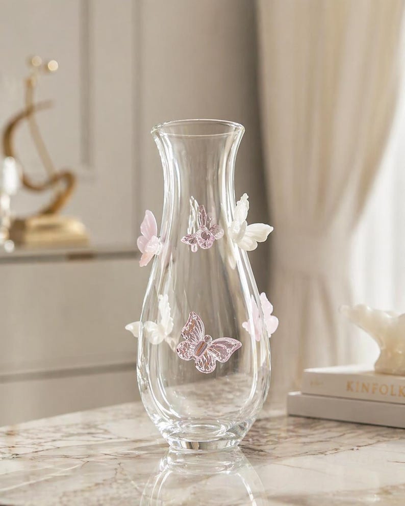 May include: A clear glass vase with a flared top and a rounded body. The vase is adorned with several decorative butterflies in shades of pink and white. The vase is sitting on a reflective surface.