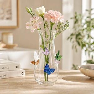 May include: A clear glass vase with colorful butterfly accents, holding a bouquet of fresh flowers. The flowers include pink carnations, white and pink alstroemeria, and white roses. The vase is on a wooden table with books and a bowl.