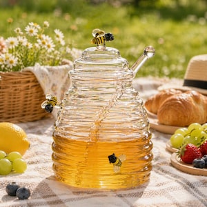May include: A clear glass honey pot with a bee-shaped lid and three decorative bees. The pot is filled with golden honey and has a glass dipper. The scene includes a picnic setting with fruit and pastries.