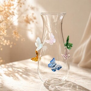 May include: A clear glass vase with a flared top, adorned with colorful glass butterflies. The butterflies are in shades of blue, green, pink, white, and gold. The vase is on a light-colored surface.