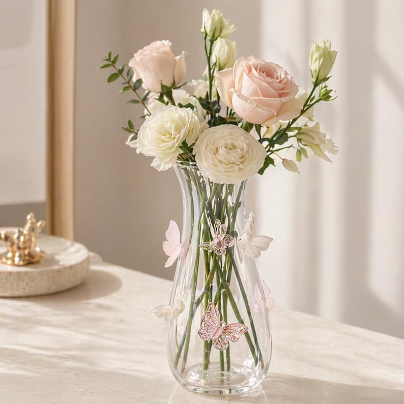May include: A clear glass vase adorned with pink and white butterflies, holding a bouquet of light pink roses and white flowers. The vase has a flared shape and is filled with water, showcasing the stems of the flowers.