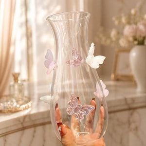 May include: A clear glass vase with a flared top and a rounded body. The vase is adorned with several translucent, pastel-colored butterfly embellishments. The butterflies are in shades of pink, white, and lavender. The vase is held by a person.