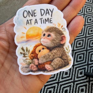 Op de afbeelding: Een sticker met twee cartoonapen die elkaar omhelzen, met een zonsondergang op de achtergrond. De tekst "ONE DAY AT A TIME" staat erboven gedrukt. De sticker wordt in een hand gehouden.