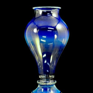May include: A tall, elegant blue glass vase with a flared top and a rounded body. The vase has a clear base and a dark blue hue that fades to lighter shades. The vase is approximately 12 inches tall.