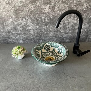 Puede incluir: Un lavabo decorativo de cerámica con un estampado floral verde y blanco. El lavabo es redondo y se encuentra sobre una superficie gris, junto a un grifo negro y una pequeña planta en maceta.