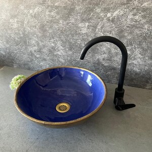 May include: A circular, blue ceramic sink with a gold-coloured rim and a brass drain. A black tap is to the right. The sink is set against a grey textured wall, suitable for a bathroom.