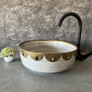 Peut inclure: Lavabo de salle de bain décoratif blanc et doré avec un robinet noir. L'évier a un bord festonné doré avec des clous décoratifs. Une petite plante en pot se trouve à gauche de l'évier.