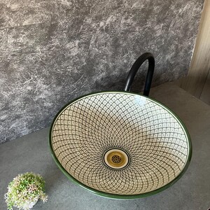 May include: Oval ceramic sink with a green rim and a cream-coloured interior featuring a black geometric pattern. The sink has a gold-coloured drain and is set on a grey countertop. A black tap is visible in the background.