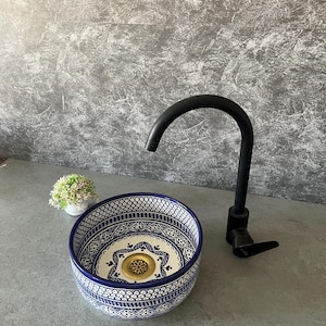 May include: A decorative ceramic sink with a blue and white patterned design. The sink is round with a gold-colored drain. A black faucet is positioned to the right of the sink. A small plant in a white pot is to the left of the sink.