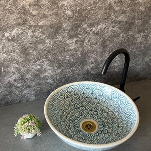 Può includere: Lavabo ovale in ceramica con un motivo a onde blu e bianche. Il lavabo ha uno scarico dorato e un rubinetto nero. A sinistra c'è un piccolo vaso bianco con della vegetazione. Un elemento decorativo per il bagno.