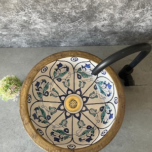 Puede incluir: Un lavabo de baño redondo y decorativo con un estampado floral en azul y verde sobre un fondo crema. El lavabo tiene un borde de madera con un borde dorado. Un grifo negro está unido al lavabo.