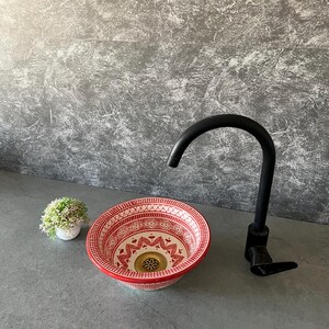 Può includere: Un lavabo in ceramica decorativo con motivi rossi e bianchi, con un rubinetto nero. Il lavabo ha un design dettagliato e un bordo rosso. Una piccola pianta in vaso è a sinistra del lavabo.