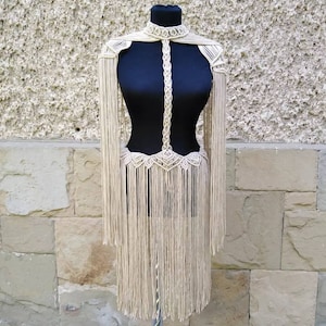 Puede incluir: Un top de macramé de color crema con detalles de flecos. El top presenta un escote halter y un corpiño ajustado.