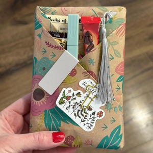 Può includere: Un regalo avvolto in carta a motivi floreali, tenuto da una persona. Il pacco contiene caffè, un pennarello azzurro e un tubetto rosso. È attaccato un adesivo con illustrazioni floreali e la scritta "cozy in the wild you".
