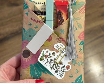 May include: A gift wrapped in floral patterned paper, held by a person. The package contains coffee, a light blue marker, and a red tube. A sticker with floral illustrations and the text "cozy in the wild you" is attached.