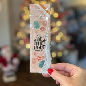 Peut inclure: Marque-page avec le texte "JUST ONE MORE Chapter" et "BE THE LIGHT Matthew 5:14". Le marque-page présente un design rose et orange avec des motifs floraux et des feuilles, et une gland argenté.