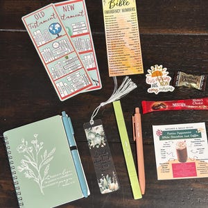 Può includere: Un assortimento di cancelleria e piccoli regali, tra cui un taccuino con design floreale, penne, segnalibri e una scheda dei numeri di emergenza della Bibbia. Gli articoli sono disposti su una superficie di legno scuro.