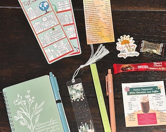 May include: An assortment of stationery and small gifts, including a notebook with floral design, pens, bookmarks, and a Bible emergency numbers card. The items are arranged on a dark wooden surface.