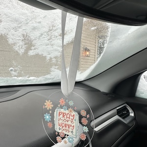 May include: A clear, circular car charm with the text "Pray more, worry less" surrounded by colorful floral designs. The charm hangs from a white ribbon, visible against a snowy backdrop.
