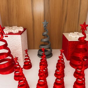 Op de afbeelding: Een verzameling decoratieve kerstbomen in rood en grijs, elk bekroond met een ster. De bomen hebben een spiraalvormig ontwerp en zijn gerangschikt op een wit oppervlak, met een houten achtergrond.