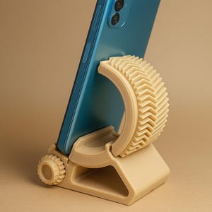 Op de afbeelding: Een turquoise smartphone staat op een beige telefoonstandaard. De standaard heeft een gebogen, getextureerde steun en een rechthoekige basis met een cirkelvormige afstelknop. De standaard is ontworpen om een telefoon rechtop te houden.