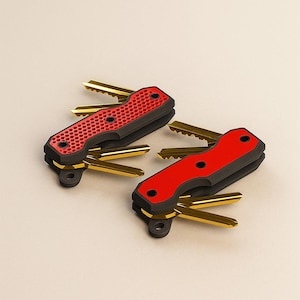 Op de afbeelding: Twee sleutelorganizers met rode en zwarte accenten. Eén heeft een gestructureerd oppervlak, de andere is glad. Beide hebben goudkleurige sleutels die zich uitstrekken vanaf de zijkanten. De sleutelorganizers staan op een neutrale achtergrond.