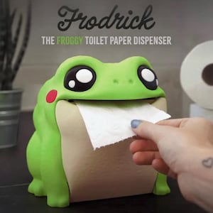May include: A green frog-shaped toilet paper dispenser with large black and white eyes and red cheek spots. The dispenser is labeled "Frodrick" and "The Froggy Toilet Paper Dispenser". A hand is pulling a sheet of white toilet paper from the frog's mouth.
