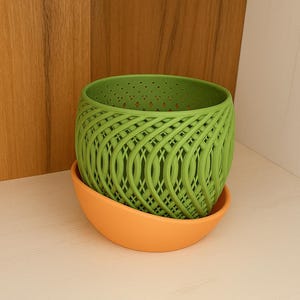 Op de afbeelding: Een groene plantenpot in geweven stijl met een oranje schotel. De pot heeft een rasterontwerp en is gemaakt van een heldergroen materiaal. De schotel is effen oranje van kleur. De plantenpot staat op een wit oppervlak.