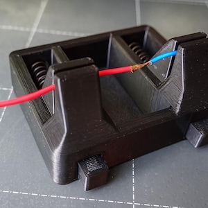 Op de afbeelding: Een zwart plastic elektrisch component met rode en blauwe draden die eruit steken. Het component heeft een rechthoekige vorm met interne compartimenten en veren. De draden zijn aan de uiteinden gestript, waardoor de koperen strengen zichtbaar worden.