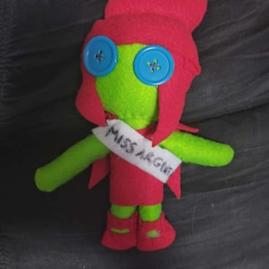 May include: A handmade felt doll with a green face and arms, red hair and dress, and blue button eyes. The doll has a white sash that reads "MISS ARGENT". The doll is a whimsical, handcrafted item.