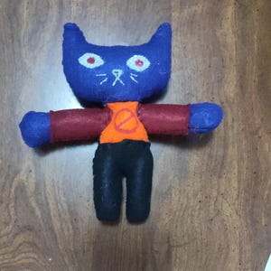 May include: A handmade plush cat doll. The doll has a blue head with red eyes and white whiskers. It wears a red shirt, an orange vest with a red circle and slash, and black trousers. The arms and feet are blue.