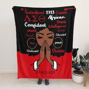Op de afbeelding: Een zwart-rode deken met een afbeelding van een vrouw met gesloten ogen en gevouwen handen. De deken bevat de woorden "Sisterhood 1913 Cream", "Confident", "smart", "loved", "Diva", "Dedicated", "Divine" en "Malcolm".