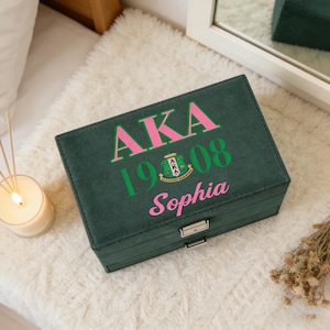 Pode incluir: Uma caixa de joias verde escuro com o texto "AKA 1908 Sophia" em rosa e verde. A caixa tem um fecho prateado e está sobre uma superfície branca e fofa. Uma vela acesa e flores secas estão no fundo.